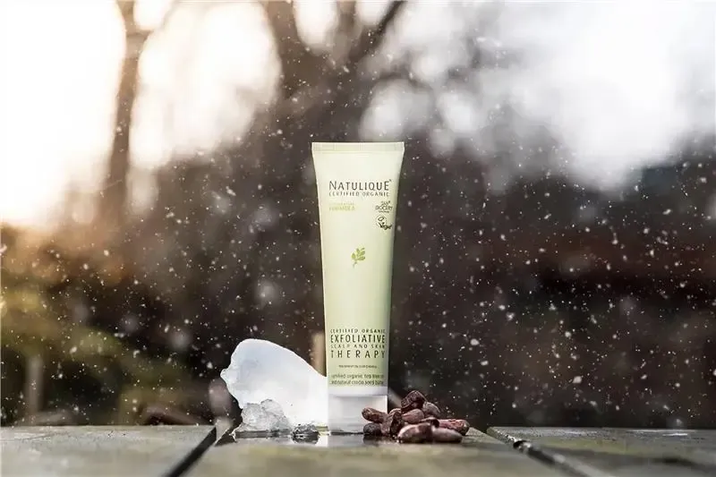 NATULIQUE Exfoliative Scalp and Skin Therapy tube on a wooden table in falling snow with ice and cocoa beans