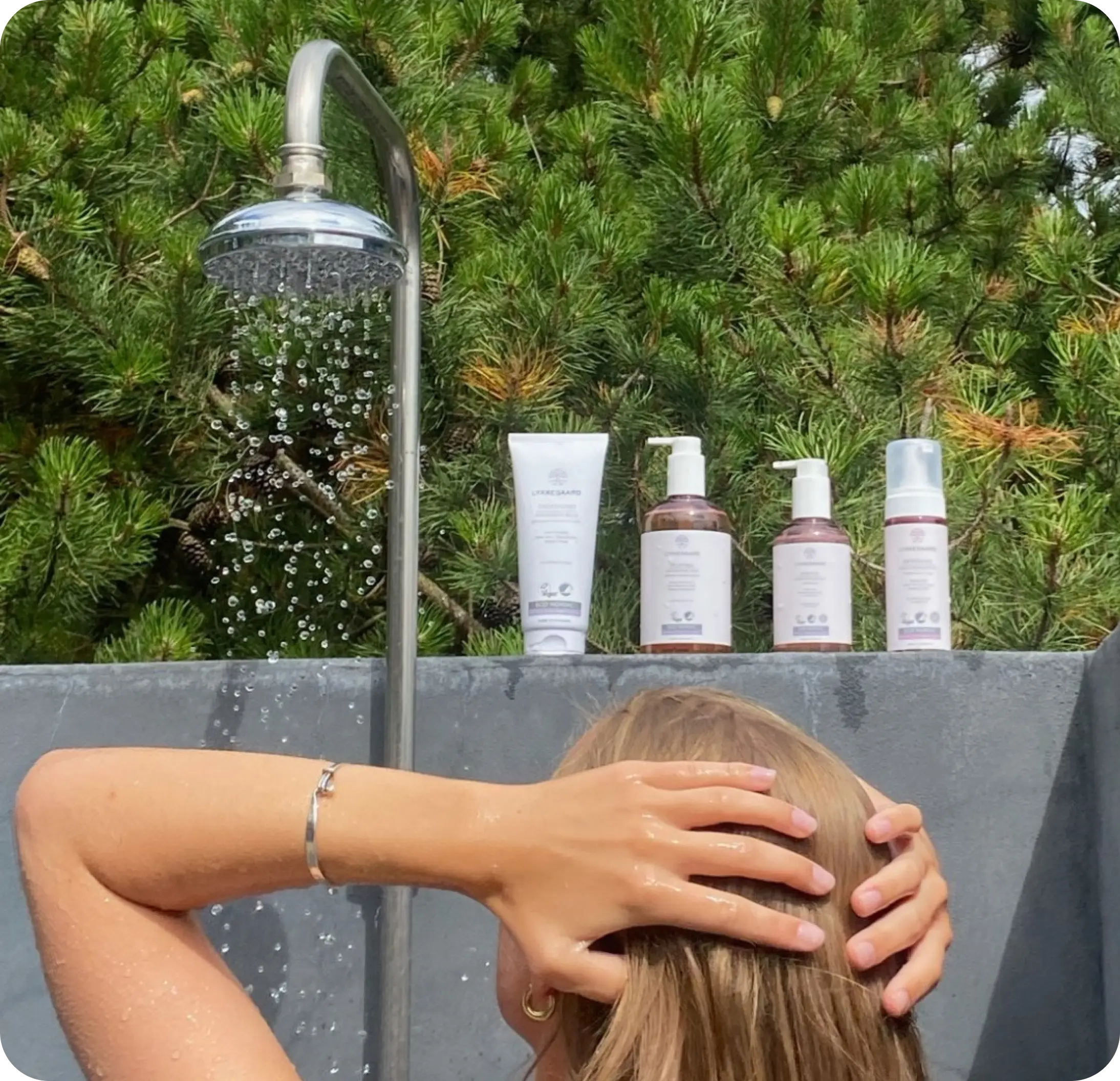 Someone rinsing their hair at an outdoor garden shower beside LYKKEGAARD skincare bottles